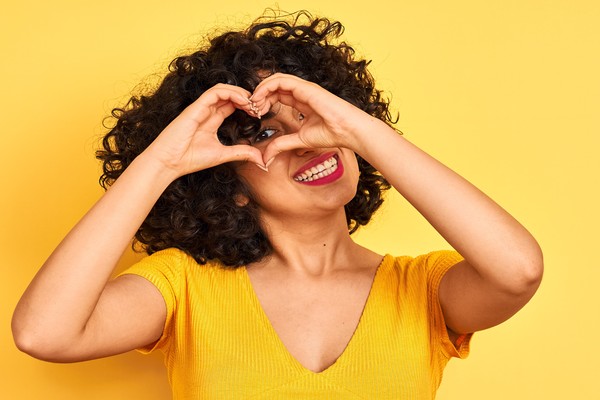Young arab woman with curly hair wearing t-shirt standing over isolated yellow background Doing heart shape with hand and fingers smiling looking through sign